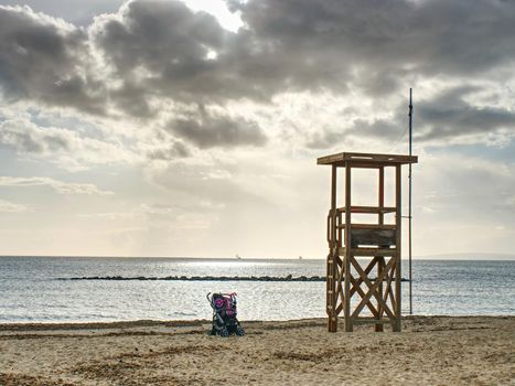 Baby carriage standing under lifeguard wooden tower on empty afternoon sand beach, calm bay level in background.