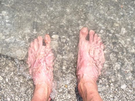 Legs and feet on the white stones of the beach with the lake or sea water. Sun reflection and mirroring in the background.