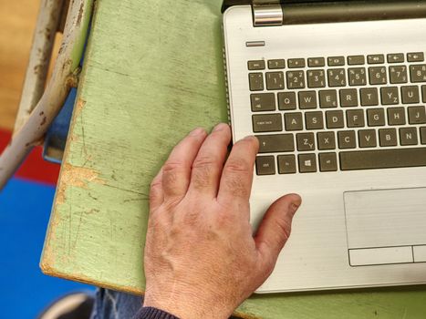 Old male freelancer typing messages via email using laptop computer and mobile phone for distant work while sitting at  worn out desk.