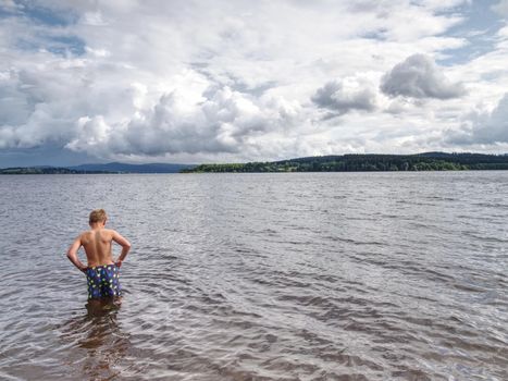 Little joyful Caucasian funny boy try cold water of lake.  Kid stay in water with level at knee 