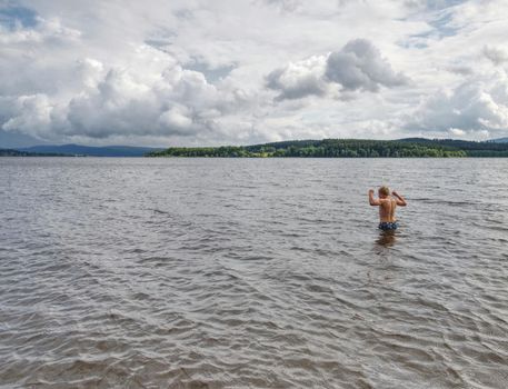 Young boy in blue swimming shorts stay in cold dark water of mountain lake. Cold weather for swimming.