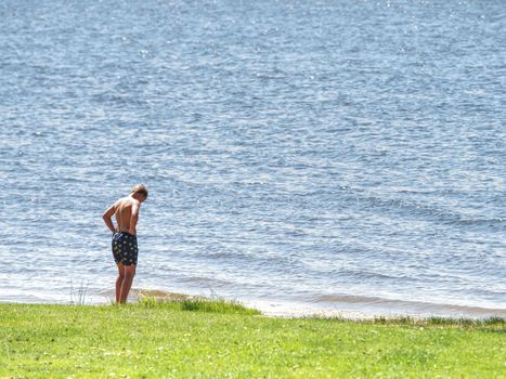 Young boy in blue swimming shorts stay in cold dark water of mountain lake. Cold weather for swimming.