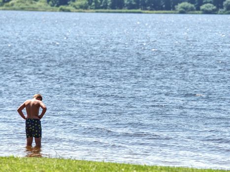 Little joyful Caucasian funny boy try cold water of lake.  Kid stay in water with level at knee 