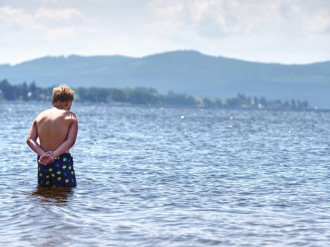 Kid stay in cold spring lake ant test cold water in sunny weather. Rear view to muscle body of boy. 