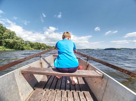 Young teenage boy rowing a rowboat or paddle boat on a lake with forest at bank. Blue summer sky in the background