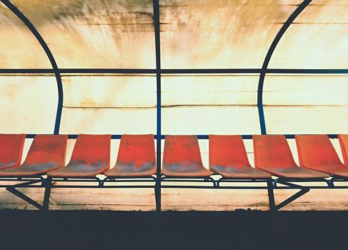 Vintage plastic seats on outdoor stadium players bench, chairs with worn paint below yellow roof.