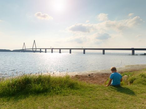 Blond hair boy is playing on sea side. Big traffic bridge between islands at the horizon. Hot summer day. 