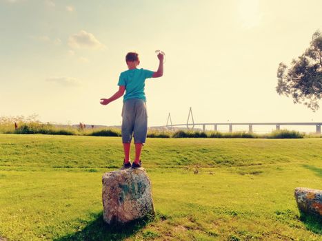 Blond hair boy is playing on sea side. Big traffic bridge between islands at the horizon. Hot summer day. 