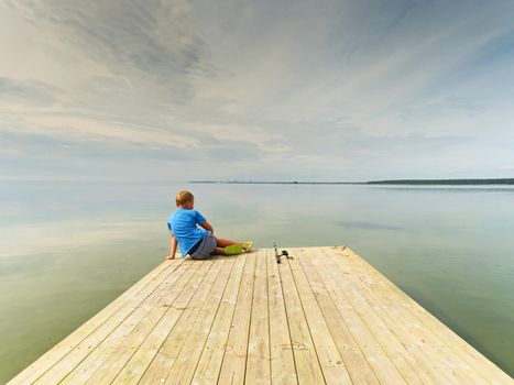 Small  fisher  on mole in summer day. Blond hair boy in blue t-shirt, grey shorts and green flip-flops is fishing on wooden board. 