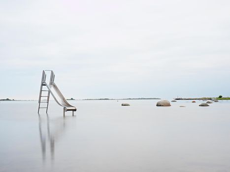 Sunset shore and old steel slide in lake. Chrome ladder tower with  sliding track, big granite stones around. Cold autumn morning with smooth water level and island at horizon.
