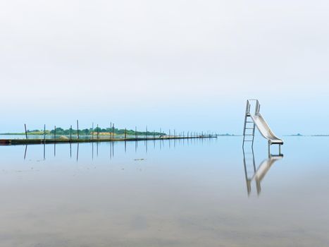 Sunset shore and old steel slide in lake. Chrome ladder tower with  sliding track, big granite stones around.  End of season in  natural swimming resort