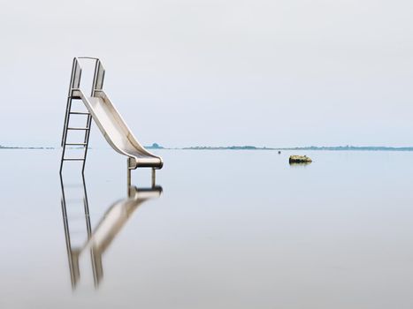 Old steel slide in lake. Chrome ladder tower with  sliding track, big granite stones around. Smooth water level and island at horizon. End of season in  natural swimming resort