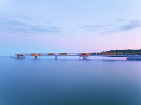 Long wooden sea bridge on coast during morning. Cold weather with first sun rays at horizon, smooth water. New built  pier in summer holiday resort. Empty mole.
