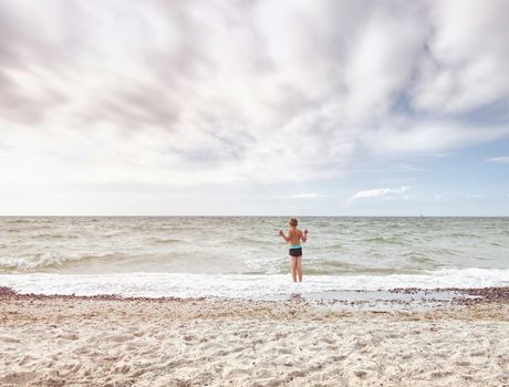 Blond hair boy stay in cold sea tide. Kid on stony beach with foamy waves.  Windy day, cloudy  blue sky on seascape 