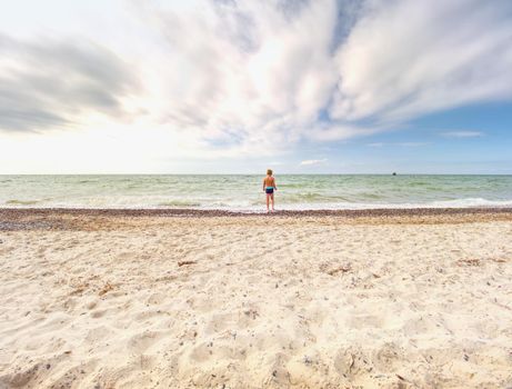 Cute fair-haired blond boy walking on beach into foamy sea waves.  Windy summer day, cloudy  blue sky on seascape background