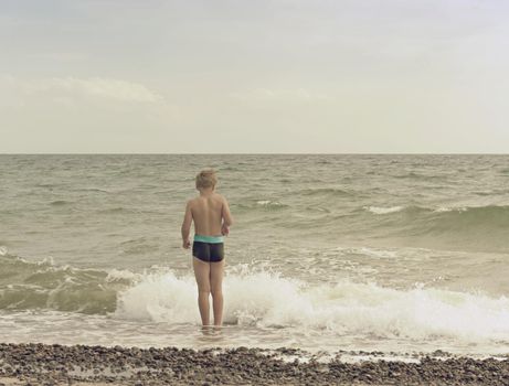 Blond hair boy stay in cold sea tide. Kid on stony beach with foamy waves.  Windy day, cloudy  blue sky on seascape 