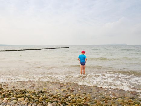 Boy in blue black sporty clothes stay in cold foamy sea. Blond hair  kid in  waves at stony beach.  Windy day, cloudy  blue sky on seascape 