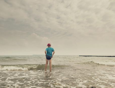 A thoughtful blond boy staying on beach near water.  Long stony beach with wooden breakwater to protect a coast  from the force of sea waves.