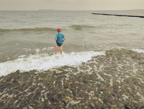 Boy in blue black sporty clothes is going into cold  water of sea. Windy day with foamy wavy sea. Blond hair  kid on stony beach.