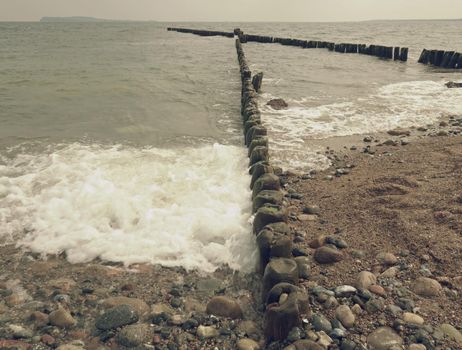 Old wooden breakwaters on a shore of Baltic Sea. White foamy water splashing stones on beach.