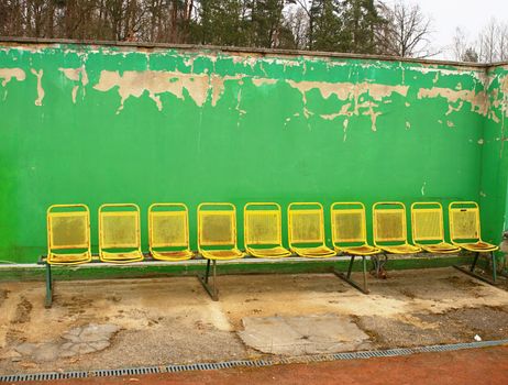 Vintage steel seats on outdoor stadium players bench chairs with worn paint below green wall