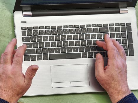 Old male freelancer typing messages via email using laptop computer and mobile phone for distant work while sitting at  worn out desk.