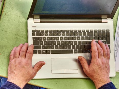 Old male freelancer typing messages via email using laptop computer and mobile phone for distant work while sitting at  worn out desk.