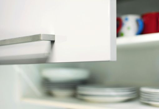 Closeup of an opened grey kitchen cabinet door with metal handle and plates and mugs inside.