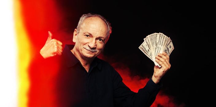 Senior gentleman holding a stack of money. Portrait of an excited old businessman. Happy old man holding dollar banknotes on dark red background with light leak. 