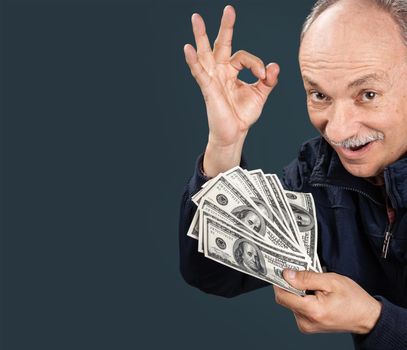 Happy elderly man showing fan of money and sign OK with fingers on dark blue background with copy-space