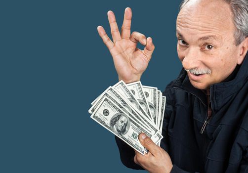 Happy elderly man showing fan of money and sign OK with fingers on blue background with copy-space