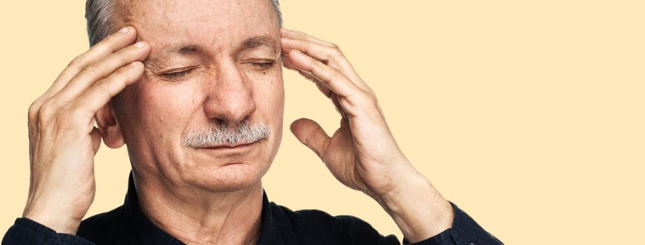 Strong headache. Old man touches his head with his hands and feeling tired and headache. Health care concept. Old man suffering from a headache isolated on yellow background with copy space.