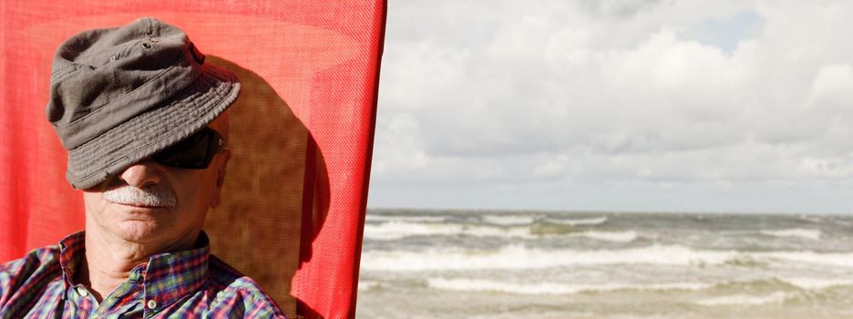 An elderly man sitting on a beach chair on the sea background covering his face by a cap. Lifestyle concept