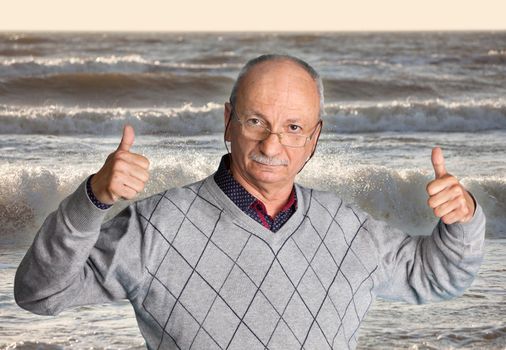 Free happy retired senior man with glasses enjoying the outdoors with wavy sea and shows thumb up.