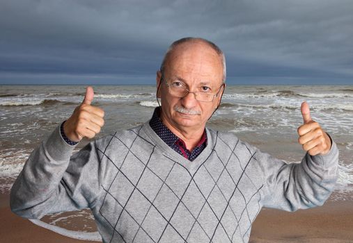Free happy retired senior man with glasses enjoying the outdoors with wavy sea and shows thumb up.