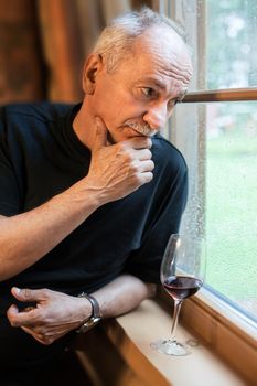 An elderly man standing near the window with a glass of wine