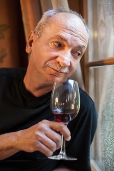 An elderly man standing near the window with a glass of wine