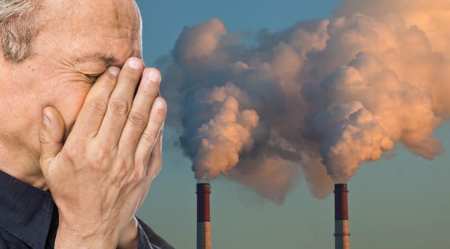 Ecological concept. Elderly man with a face closed by hands against the background of pipes polluting an atmosphere
