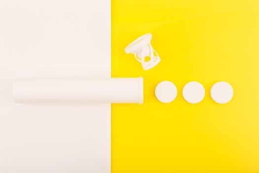 Creative simple flat lay with white opened tube with spilled effervescent vitamins on white and yellow background. Concept of vitamin C, supplements and healthcare