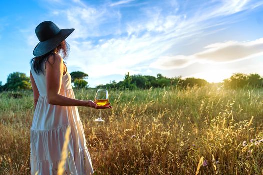 Unrecognizable lonely woman in boho white long dress with large dark hat, looking to the sunset in a wheat field holding a white wine glasses in hand. Concept of dream life in contact with nature