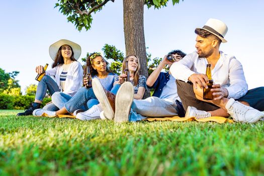 Group of five happy young diverse multiracial gen z people in outdoor party drinking beer from bottle talking each other sitting on grass of city park meadow. Friends having fun with alcohol outdoor
