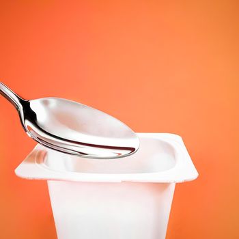 Yogurt cup and silver spoon on orange background, white plastic container with yoghurt cream, fresh dairy product for healthy diet and nutrition balance.