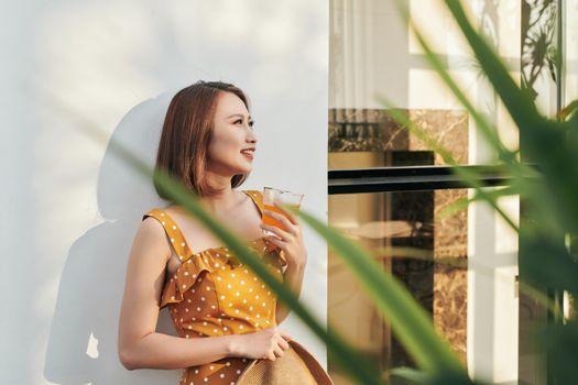 Pretty Asian girl wearing vintage dress and holding hat with orange juice behind wall