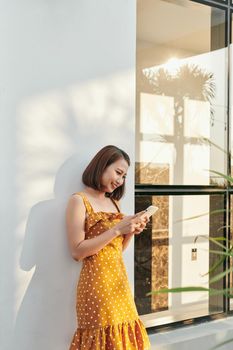Portrait of young smiling fashion girl standing the wall and using smartphone