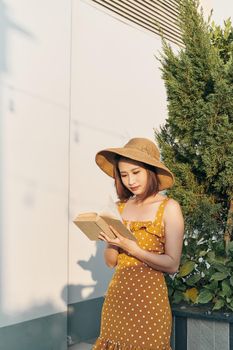 Young woman reading a book standing lean against trunk tree in summer park outdoor.