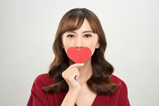 Head and shoulders portrait of attractive Asian woman looking at camera with deep gray eyes while covering her mouth with heart shaped piece of paper