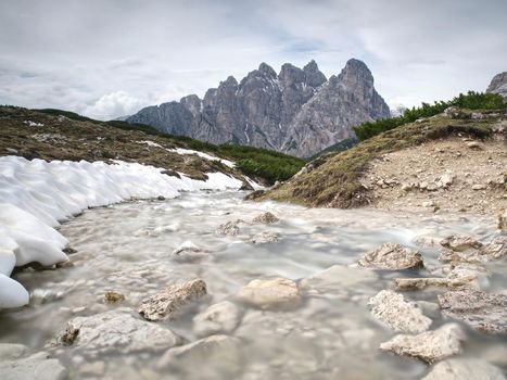 Mountains landscape wild stream in front. White snowy mountain peaks and grey cloudy sky