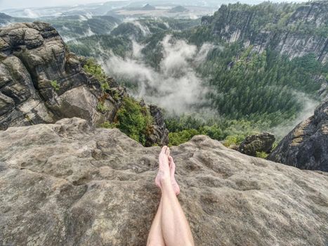 The crossed legs take a rest on tiring mountain trail. Sweaty male legs without trousers relaxing on peak of mountain above valley.