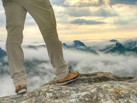 Hiker legs in light outdoor trousers and comfortable leather  trekking boots on rocky peak. Misty valley below peak in far blurry background. Hiking in nature.