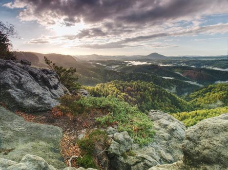 Peak of sandstone rock with view into colorful mist in morning valley. Blue eyllow daybreak sun hidden at horizon